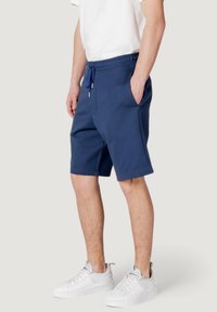 Pantalones cortos de chándal azul marino hechos de tejido suave, con una cinturilla elástica y cordón, bolsillos laterales y un corte recto. Se llevan zapatillas blancas.