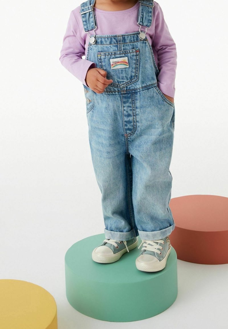 Enfant portant une salopette en jean, un t-shirt à manches longues violet et des baskets, debout sur une plate-forme ronde verte avec des plateformes jaunes et marron à proximité.
