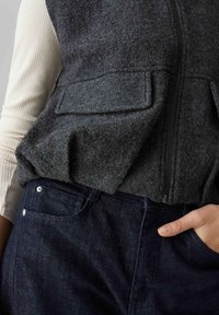 Gilet di feltro grigio con cerniera e due tasche sul petto, abbinato a pantaloni in denim scuro con tasca e dettagli in rivetto metallico.