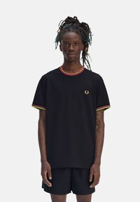 Camiseta negra con cuello redondo, adornada con ribetes rojos, amarillos y verdes, y un emblema dorado en el pecho. Diseño simple, de manga corta.