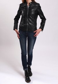 Černá kožená bunda s přiléhavým střihem, stojáčkovým límcem a zapínáním na zip, nošená přes modré džíny a černé šněrovací boty.