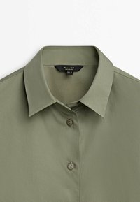 Chemise vert olive clair à col avec deux boutons visibles et une étiquette de marque noire à l'intérieur de l'encolure.