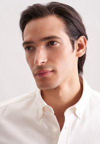 Jonge man met donker haar in een wit blinkshirt, die nadenkend opzij kijkt tegen een effen lichte achtergrond.