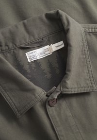 Camisa de botones verde oliva con cuello estructurado y tela texturizada. Cuenta con una etiqueta con un patrón de bosque e instrucciones de cuidado.