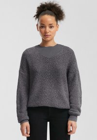 Grijze gebreide trui met een V-hals, brede mouwen en een textuurpatroon. Gestyled over een donkergrijze top, gecombineerd met zwarte broeken.
