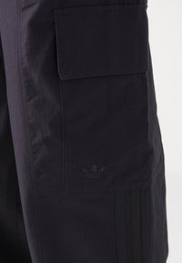 Tmavě fialové cargo šortky s vertikálními pruhy, se postranní kapsou a texturovaným logem Adidas. Obsahují černé postranní pruhy.