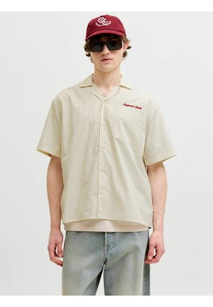 Jeune homme portant une chemise beige à fines rayures manches courtes avec un texte rouge, un jean bleu clair, une casquette bordeaux et de grandes lunettes de soleil noires sur un fond uni.