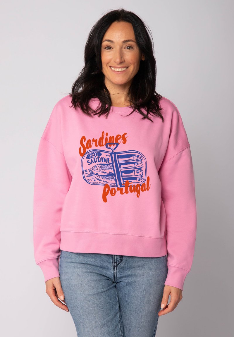 Eine lächelnde Frau trägt einen rosa Sweatshirt mit einem blauen Sardinen-Dose-Graphic und orangem Text "Sardinen Portugal", kombiniert mit hellblauen Jeans.