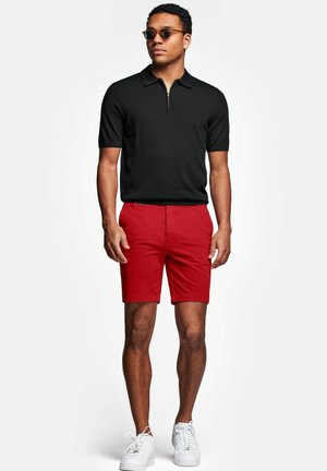 Homme portant un t-shirt noir à manches courtes avec col zippé, un short rouge, des baskets blanches et des lunettes de soleil, debout devant un fond blanc uni.