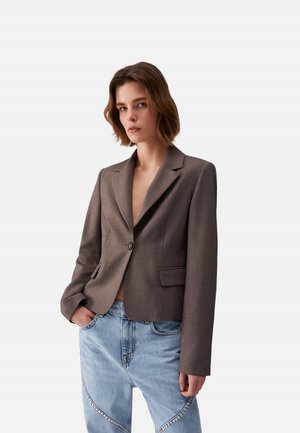 Giovane che indossa un blazer marrone aderente con un bottone e jeans azzurri con cuciture decorative, in piedi con una mano in tasca.