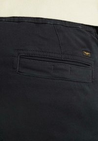 Schwarze Hose mit einer Gesäßtasche, die eine rechteckige Klappe, kontrastierende Nähte und ein kleines dekoratives Etikett aufweist. Glatte Stofftextur.