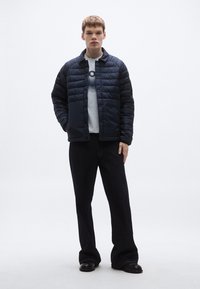 Granatowa pikowana kurtka z zapięciem na guziki, noszona na jasnoszarej koszuli i ciemnych spodniach, z teksturowanym, pikowanym wzorem.