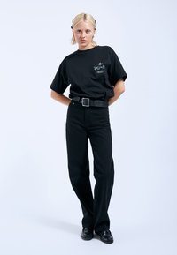 Černé oversized tričko s grafickým potiskem, spárované s černými džíny s vysokým pasem a širokým černým páskem. Model má na sobě černé boty.