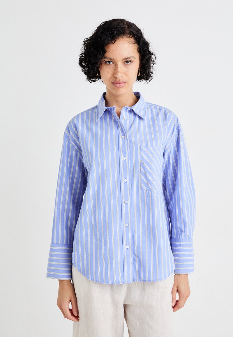 Camicia a righe blu e bianche con chiusura a bottoni, colletto e taschino singolo; presenta maniche lunghe con accenti a righe.
