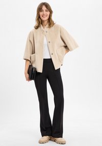 Beige kurze Jacke mit weicher Textur, ausgestattet mit zwei frontalen Taschen und drei großen Knöpfen. Kombiniert mit schwarzen engen Hosen und beigen Schuhen.