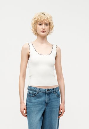 Žena s krátkymi kučeravými blond vlasmi, oblečená v bielom topu s vlnitými okrajmi bez rukávov a modrými džínami s vysokým pásom, stojaca pred jednofarebným pozadím.