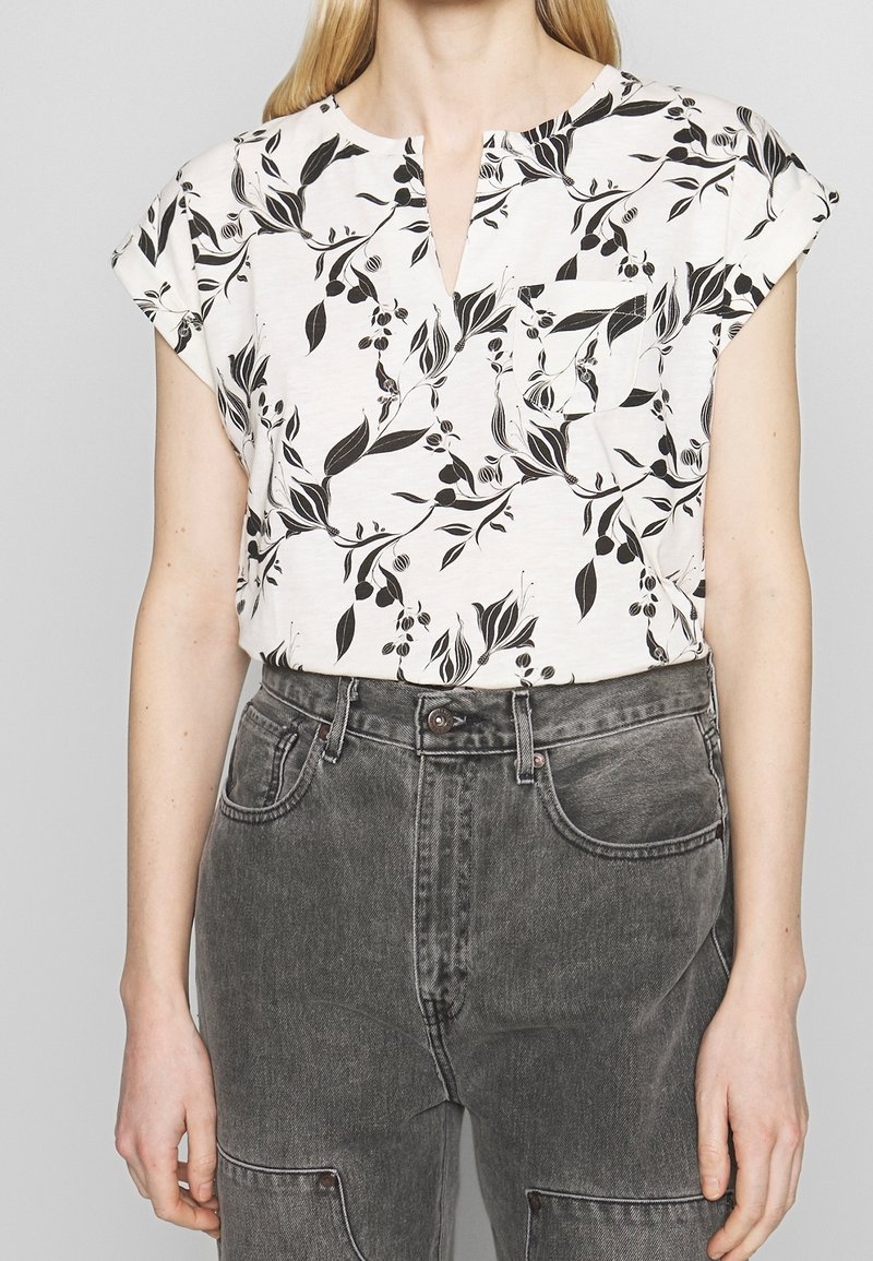 Hvid kortærmet top med sort blomster mønster, featuring en lille lomme foran og en V-hals design, parret med mørkegrå denim jeans.