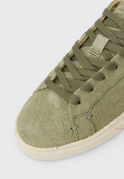 Αθλητικό παπούτσι από σουέντ σε ελιά πράσινο χρώμα με υφή στην επάνω επιφάνεια, επίπεδα κορδόνια, στρογγυλεμένη μύτη και μπεζ ελαστική σόλα. Διαθέτει διακοσμητικές ραφές.