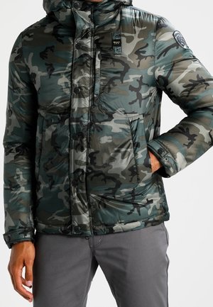 Mężczyzna w zielono-szarej pikowanej kurtce kamuflażowej z kapturem i kieszeniami po bokach, w szarych spodniach, stoi z jedną ręką w kieszeni.