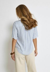 Camicia a maniche corte a righe blu chiaro e bianche con orlo curvo, yoke posteriore e vestibilità rilassata, abbinata a pantaloni beige a gamba larga.