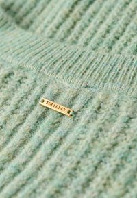 Texturerad grön tröja med ett stickat mönster och upphöjda ribbade detaljer. Guldig metalltagg med "SUPERDRY"-logotyp fäst.