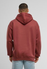 Bordová mikina s priestrannou kapucňou, spadnutými plecami a jemne rebrovanými manžetami. Hladký povrch a uvoľnený strih. Nosená s svetlomodrými džínsami.