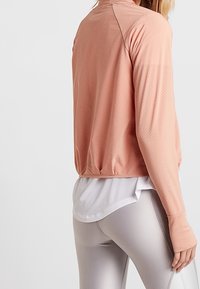 Mujer vistiendo una camiseta atlética de manga larga melocotón sobre una camiseta de malla blanca y leggings gris claro, mostrada desde la espalda y el lado.