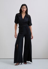 Combinaison noire avec des manches courtes bouffantes, une ceinture smockée et des fentes latérales. Présente des motifs texturés le long des jambes.