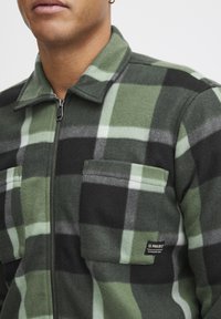 Fleecejack met een groen en zwart ruitenpatroon, ritssluiting, twee borstzakken en een label met het merk "11 PROJECT."