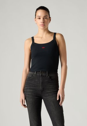 Femme portant un débardeur noir avec un petit logo rouge et un jean noir taille haute, debout devant un fond gris clair uni.