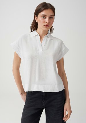Femme aux cheveux bruns et lisses portant un chemisier blanc ample à manches courtes avec col et un jean noir, debout devant un fond clair et uni.