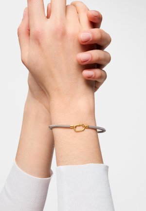 Dos manos entrelazadas con una muñeca que lleva una pulsera delgada de plata con un broche dorado de forma ovalada.