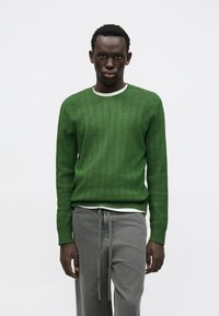 Zielony dziergany sweter o prążkowanej fakturze, z okrągłym dekoltem i długimi rękawami, noszony na białej koszuli i zestawiony ze szarymi spodniami.