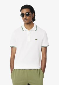 Polo shirt blanc avec des accents rayés verts sur le col et les manches, avec un petit logo brodé sur la poitrine gauche.