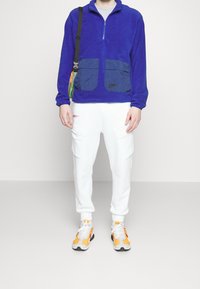 Blå fleece zip-up tröja med två navy-färgade framfickor. Vita cargobyxor med sidofickor. Flerfärgade sneakers med orange detaljer.