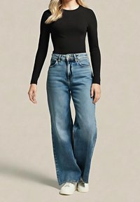 Femme portant un haut noir à manches longues, un jean bleu taille haute à jambes larges, et des baskets blanches, debout devant un fond uni.