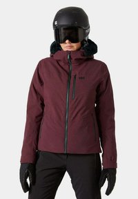 Giacca da sci rossa con zip frontale, colletto alto e accenti neri. Dotata di un morbido cappuccio foderato in pelliccia e materiale impermeabile. Gli accessori includono un casco nero e guanti.