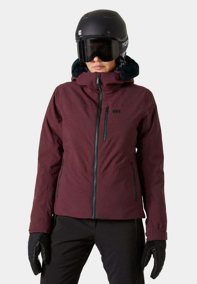 Giacca da sci rossa con zip frontale, colletto alto e accenti neri. Dotata di un morbido cappuccio foderato in pelliccia e materiale impermeabile. Gli accessori includono un casco nero e guanti.