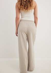 Kvinna med långt lockigt hår som bär en vit ärmlös topp och lösa beige byxor, står mot en enfärgad ljus bakgrund, sedd bakifrån.