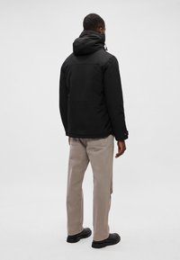 Chaqueta negra con capucha, cierre de cremallera, puños elásticos y una tela texturizada. Combinada con pantalones beige claros y zapatos negros.