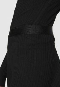 Czarna tkanina z prążkami, z gładkim, błyszczącym czarnym paskiem. Tekstura jest miękka z pionowymi liniami, podkreślającymi smukłą sylwetkę.