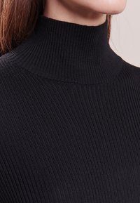 Zwarte geribbelde coltrui met een aangesloten pasvorm. De stof voelt zacht aan en de kraag heeft een hoge, aansluitende vorm.