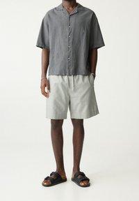 Chemise grise à manches courtes avec fermeture à boutons, short vert clair avec ceinture élastique, et sandales noires à enfiler avec une boucle décorative.