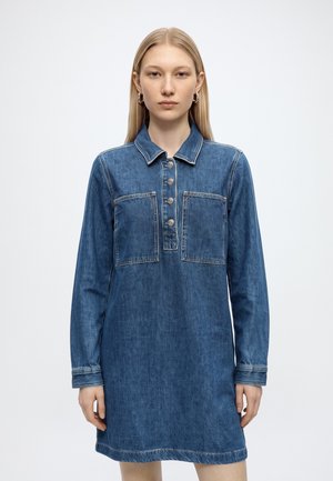 Young woman with straight blonde hair wears a blue denim dress with long sleeves, front pockets, and buttons down the chest.