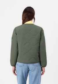 Grøn quiltet jakke med bølget mønster, gul krave og lange ærmer. Iført med lyseblå denim jeans. Glat tekstur, afslappet design.