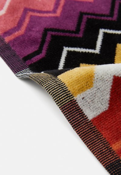 Multicolored geometric patterned rug featuring zigzag designs in purple, yellow, orange, and black, with a textured pile and striped edge.