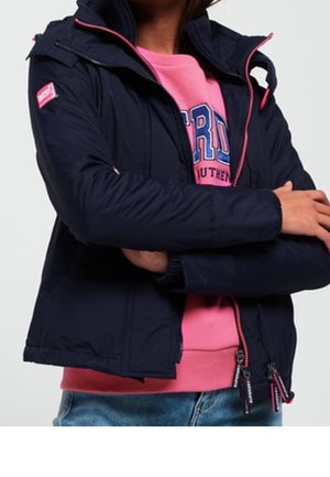 Person trägt eine dunkelblaue Jacke über einem rosa Sweatshirt mit blauer Schrift, die Arme verschränkt, und blaue Jeans sind an der Taille sichtbar.