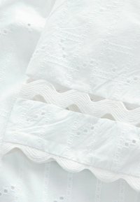 Tissu blanc avec des motifs floraux brodés et des bords festonnés créant un design texturé et décoratif.