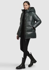 Donkergroene pufferjack met een glanzende afwerking, voorzien van een capuchon, rits en zijzakken. Gemaakt in combinatie met zwarte, strakke leggings en laarzen.