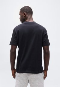 Άνδρας που στέκεται με την πλάτη στραμμένη, φοράει απλό μαύρο κοντομάνικο μπλουζάκι και ανοιχτό γκρι φόρμας, πάνω σε λευκό φόντο.